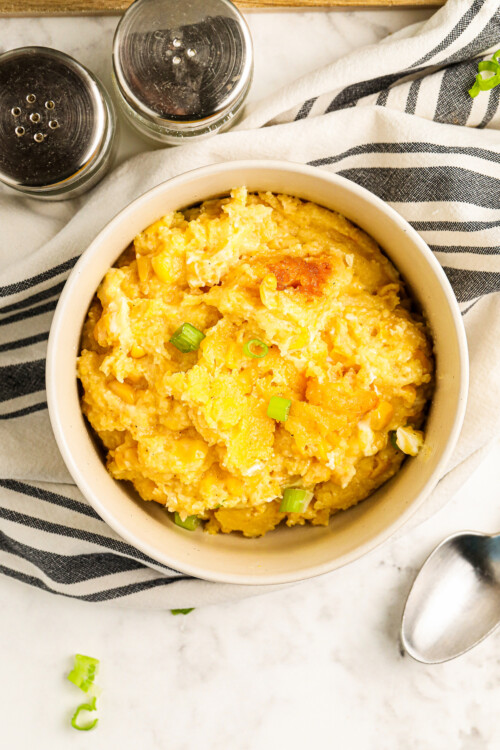 A bowl of sweet and savory crockpot corn casserole topped with sliced green onions with a spoon, salt and pepper on the side.
