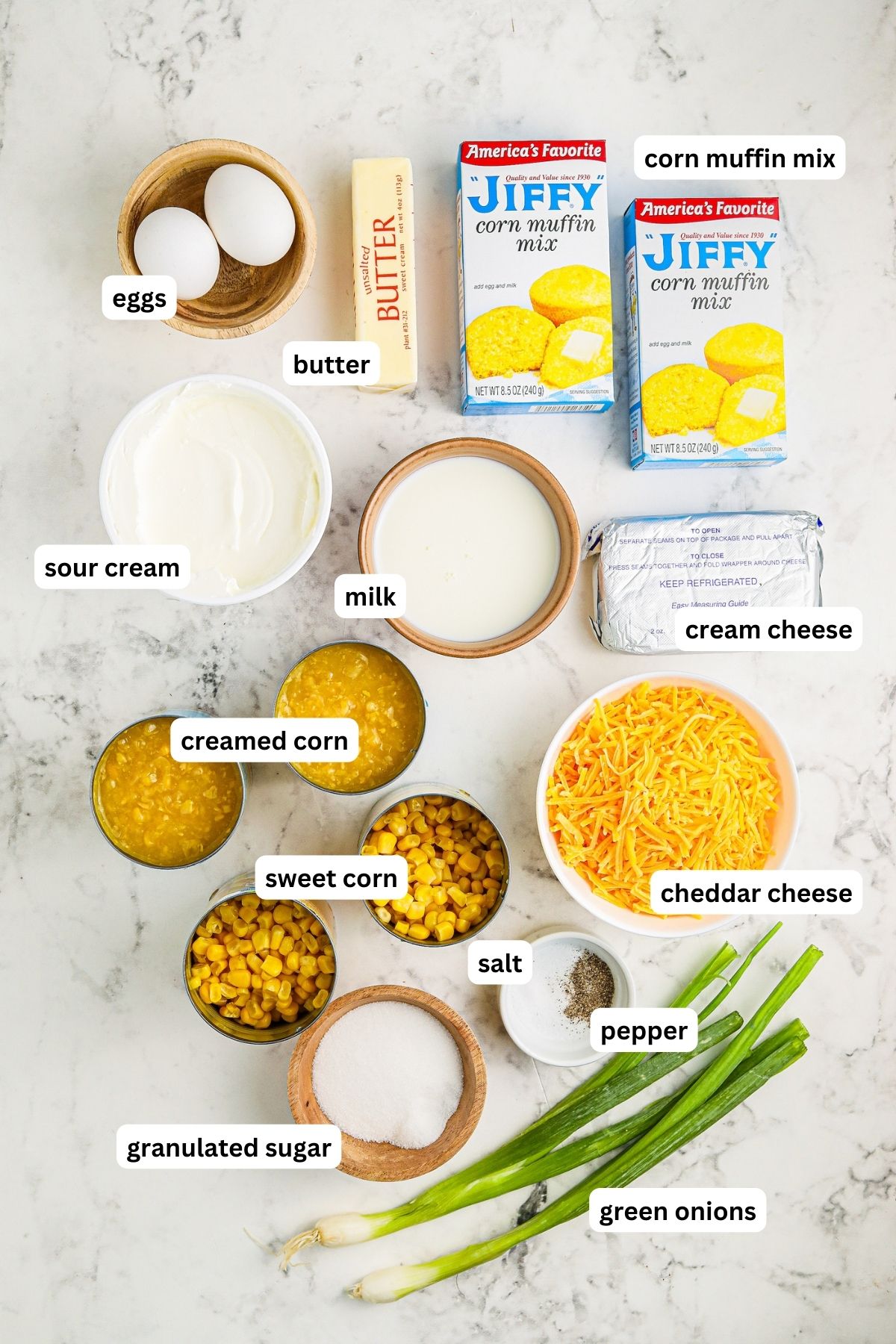 Ingredients arranged in bowls for crockpot corn pudding recipe. From top to bottom: eggs, butter, corn muffin mix, sour cream, milk, cream cheese, creamed corn, corn kernels, shredded cheddar cheese, granulated sugar, salt, pepper and green onions.
