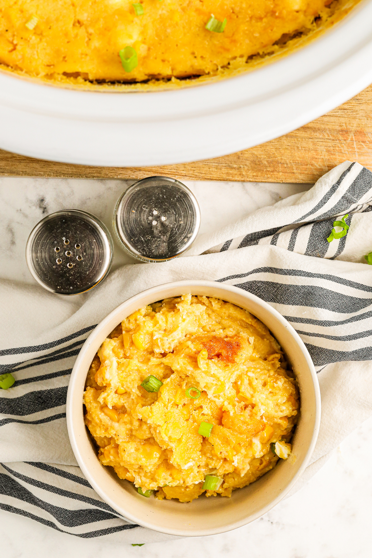 A serving of sweet and creamy crockpot corn casserole in a bowl topped with sliced green onions with a crockpot full of corn pudding in the background.