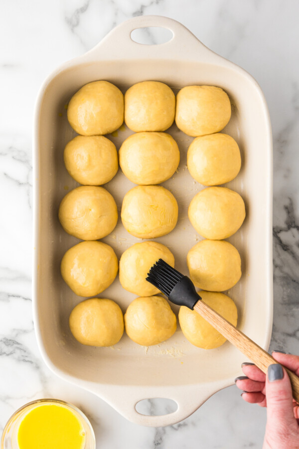 Balls of yeast dough in a baking dish being brushed with egg wash.
