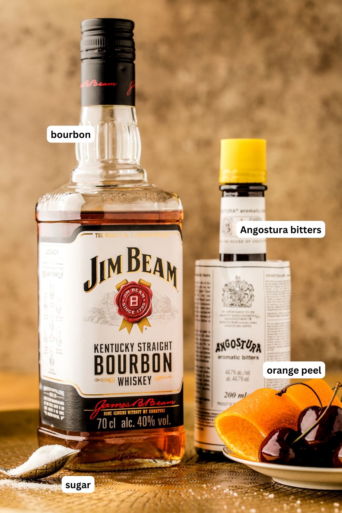 Labeled ingredients for old fashioned recipe, from top to bottom: bourbon, angostura bitters, orange peel, sugar, and optional cherries for garnish.