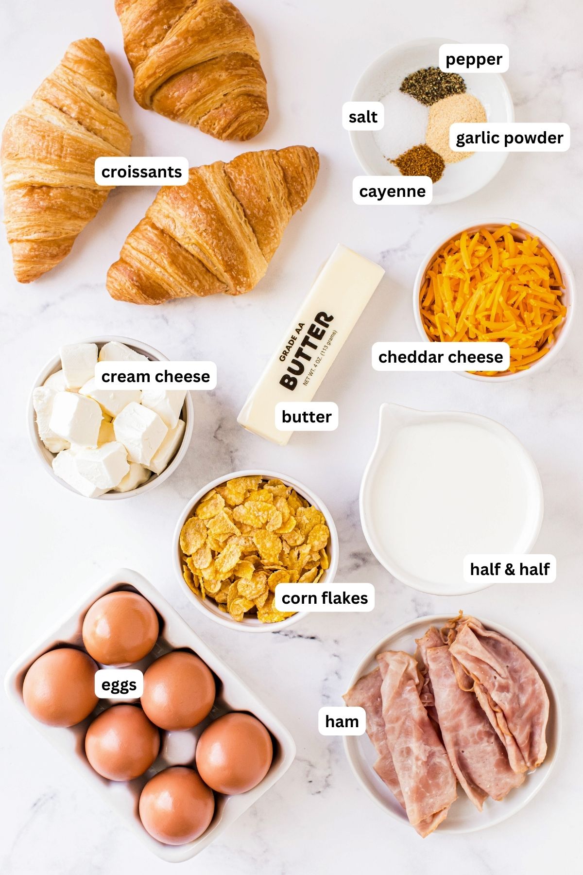 Ingredients for overnight breakfast casserole recipe arranged on a countertop. From top to bottom: croissants, pepper, salt, onion powder, cayenne pepper, cream cheese, butter, cheddar cheese, half and half, cornflakes, eggs and sliced ham.