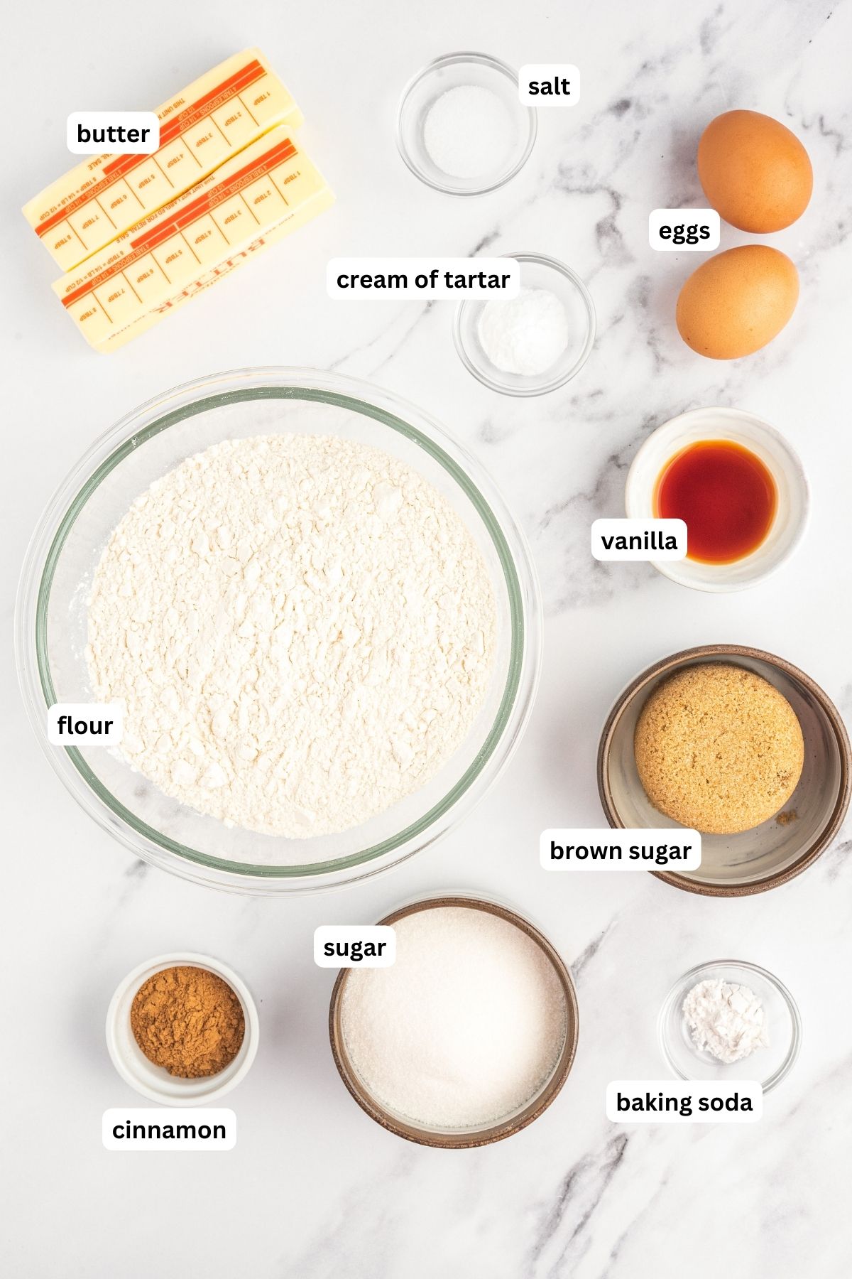 Ingredients for snickerdoodle recipe arranged in bowls, from top to bottom: salt, butter, cream of tartar, eggs, vanilla, flour, brown sugar, sugar, cinnamon and baking soda.