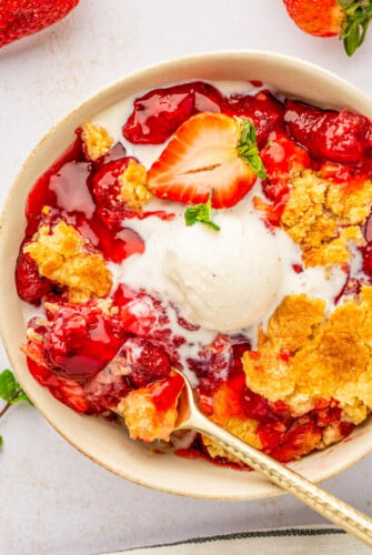 A bowl of warm strawberry dump cake with a scoop of vanilla ice cream on top with a fork taking a bite.