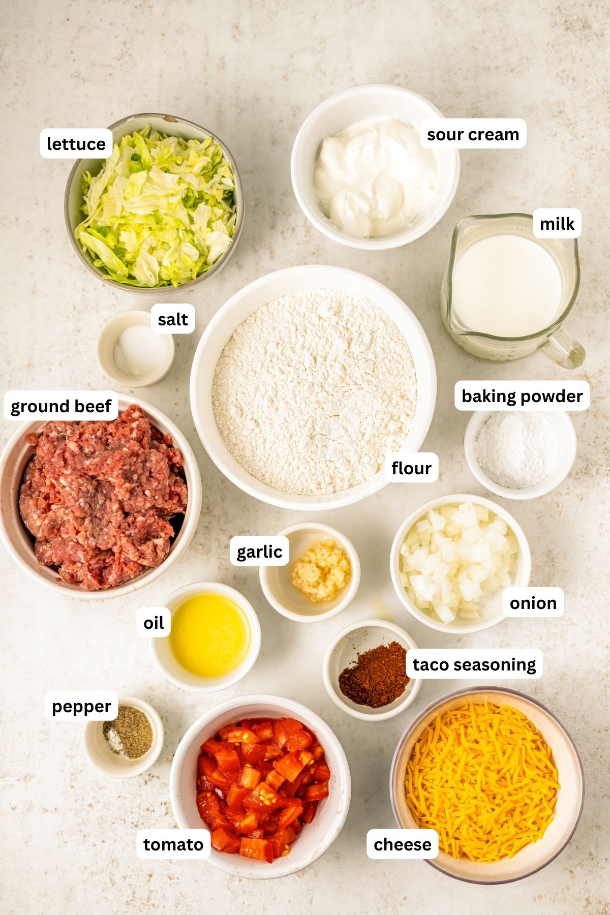 Ingredients for homemade Taco Bell chalupa recipe arranged in bowls. From top to bottom: lettuce, sour cream, milk, salt, flour, baking powder, ground beef, garlic, onion, oil, taco seasoning, pepper, tomato and shredded cheese.