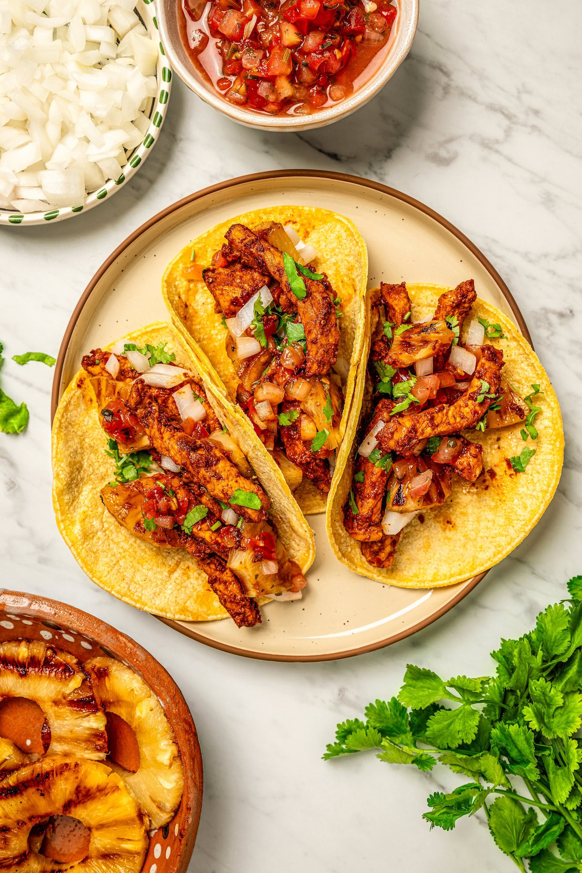 A plate of three authentic Al Pastor Tacos served on corn tortillas with grilled pineapple, diced onion and sals in bowls on the side.