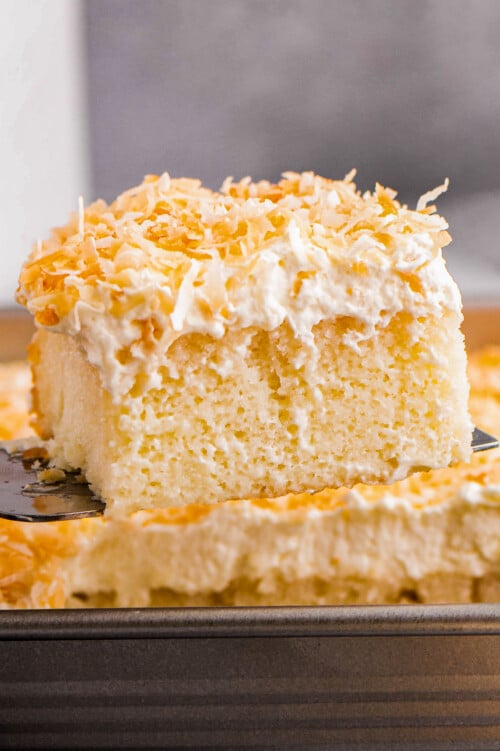A slice of moist coconut poke cake being served with a cake serve out of a baking pan.