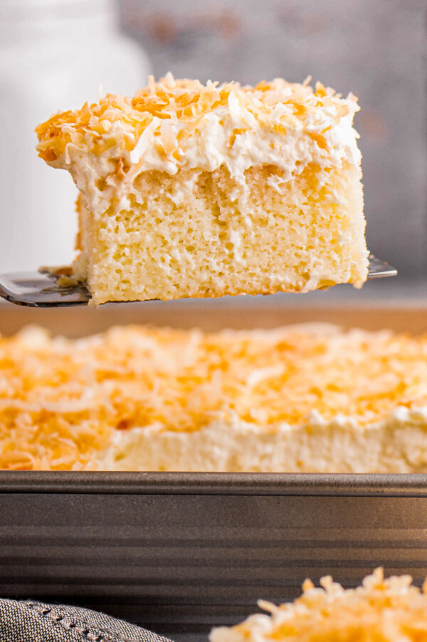 A slice of moist and creamy coconut cake topped with cream of coconut, coconut whipped cream and toasted coconut being served out of a cake pan.