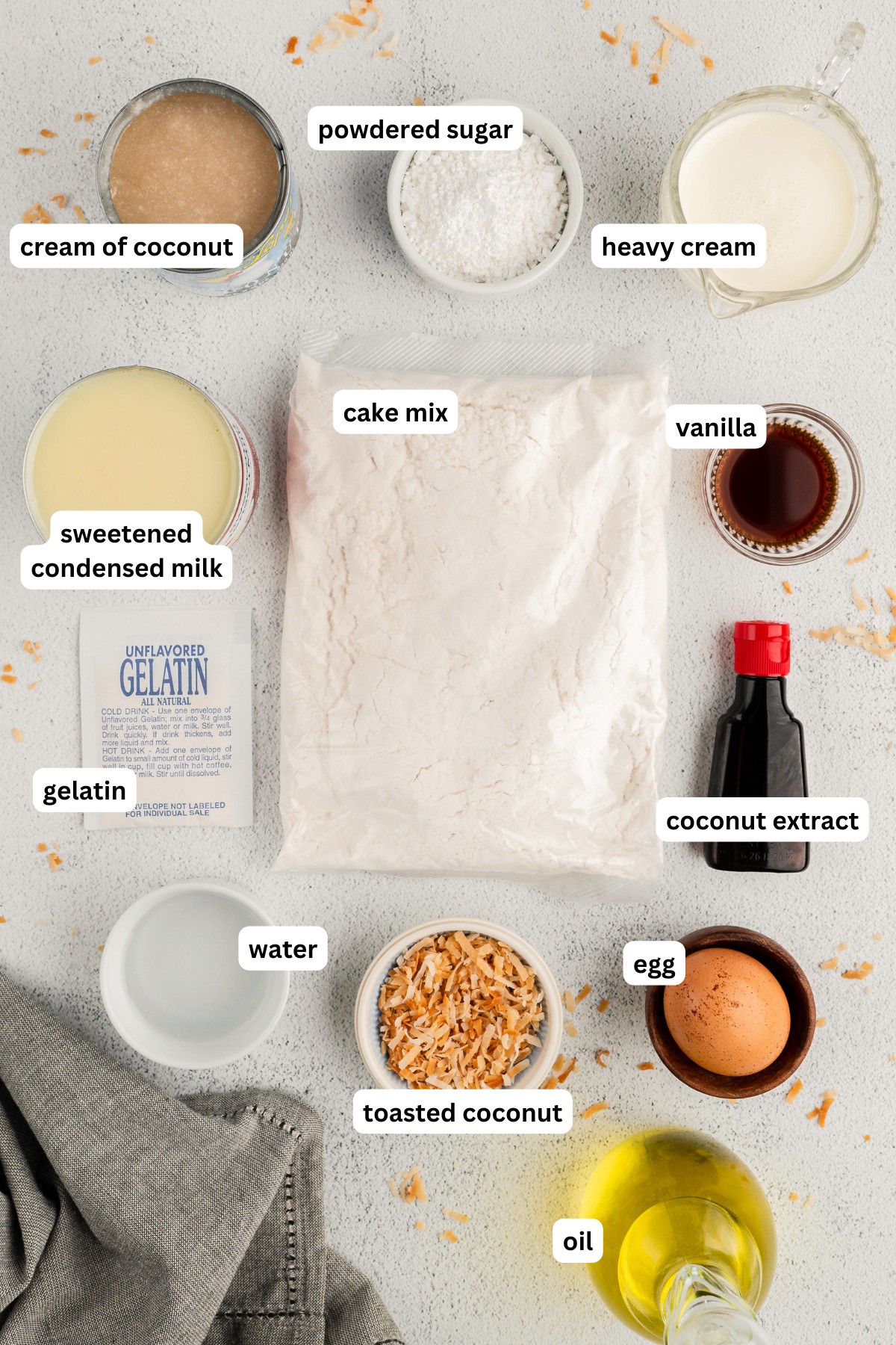 Ingredients for coconut poke cake recipe arranged on a countertop. From top to bottom: cream of coconut, powdered sugar, heavy cream, sweetened condensed milk, cake mix, vanilla extract, gelatin, coconut extract, water, toasted coconut, egg and oil.