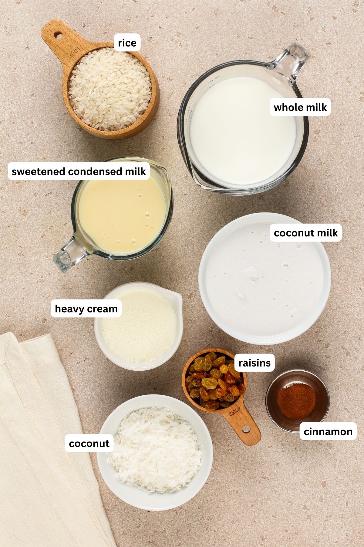 Coconut rice pudding recipe ingredients arranged in bowls. From top to bottom: rice, whole milk, sweetened condensed milk, coconut milk, heavy cream, raisins, cinnamon and shredded coconut.