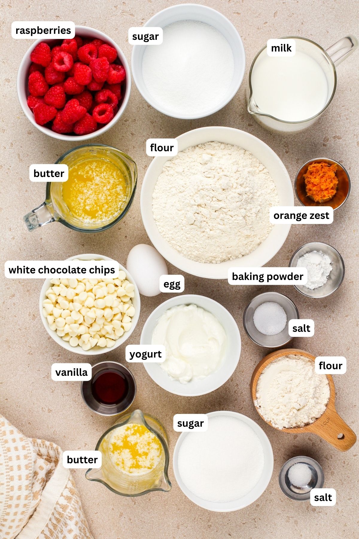 Raspberry muffin recipe ingredients arranged in bowls, from top to bottom: raspberries, granulated sugar, milk, butter, flour, orange zest, white chocolate chips, egg, baking powder, salt, yogurt, and vanilla extract.