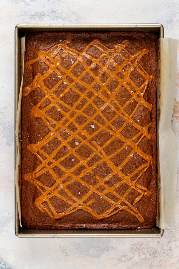 Baked caramel and chocolate brownies with flaky sea salt on top in a baking pan lined with parchment paper.