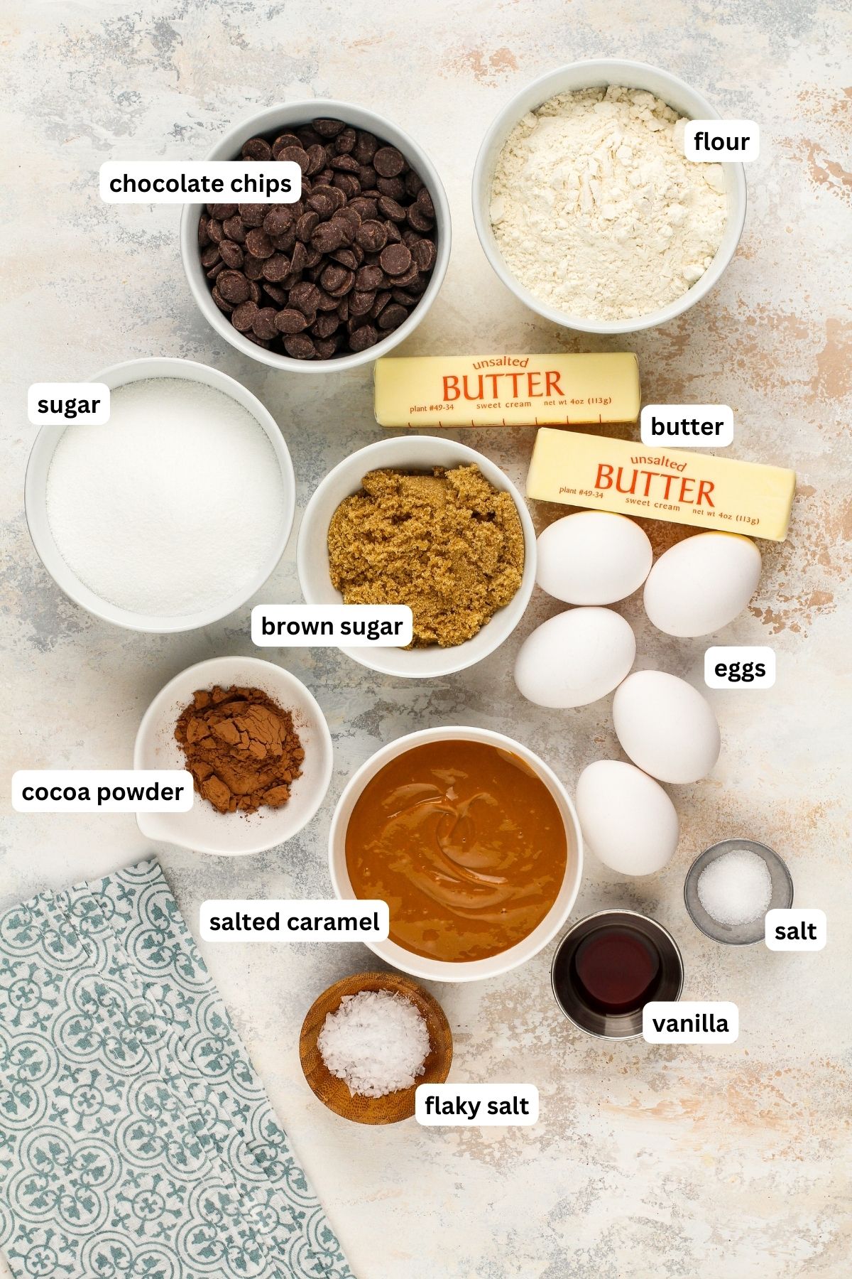 Salted caramel brownies recipe ingredients arranged in bowls. From top to bottom, chocolate chips, flour, sugar, butter, brown sugar, eggs, cocoa powder, salted caramel sauce, salt, vanilla and flaky sea salt.