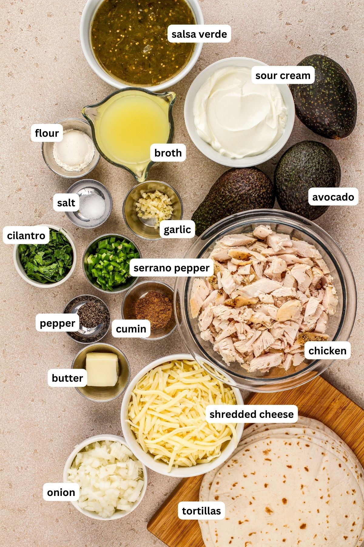 Ingredients for white chicken enchilada recipe arranged in bowls. From top to bottom: salsa verde, sour cream, flour, broth, avocado, salt, garlic, cilantro, serrano pepper, cooked chicken, pepper, cumin, butter, shredded cheese, onion and tortillas.
