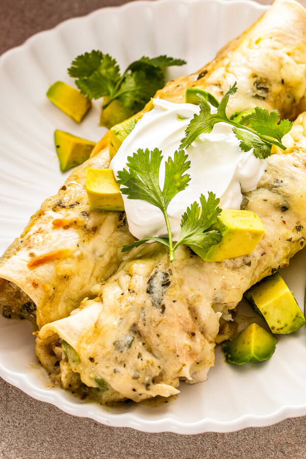 A serving of creamy chicken enchiladas with white sauce and lots of gooey cheese topped with sour cream, diced avocado and fresh cilantro.