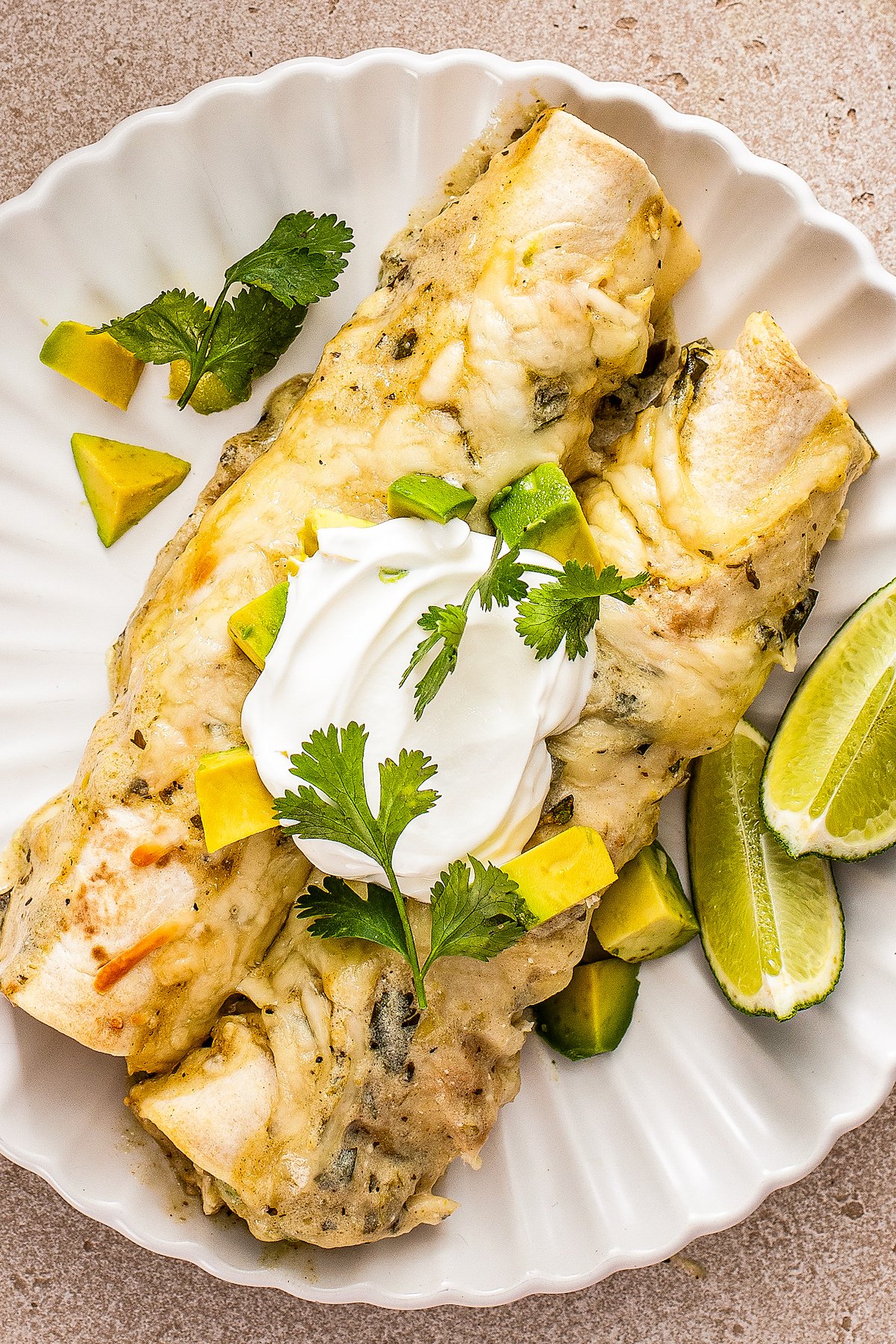 A plate of two creamy chicken enchiladas topped with sour cream, diced fresh avocado and cilantro.