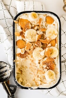 A container of creamy banana pudding with vanilla wafers and sliced banana on top with a scoop of ice cream missing and an ice cream scoop on the side.