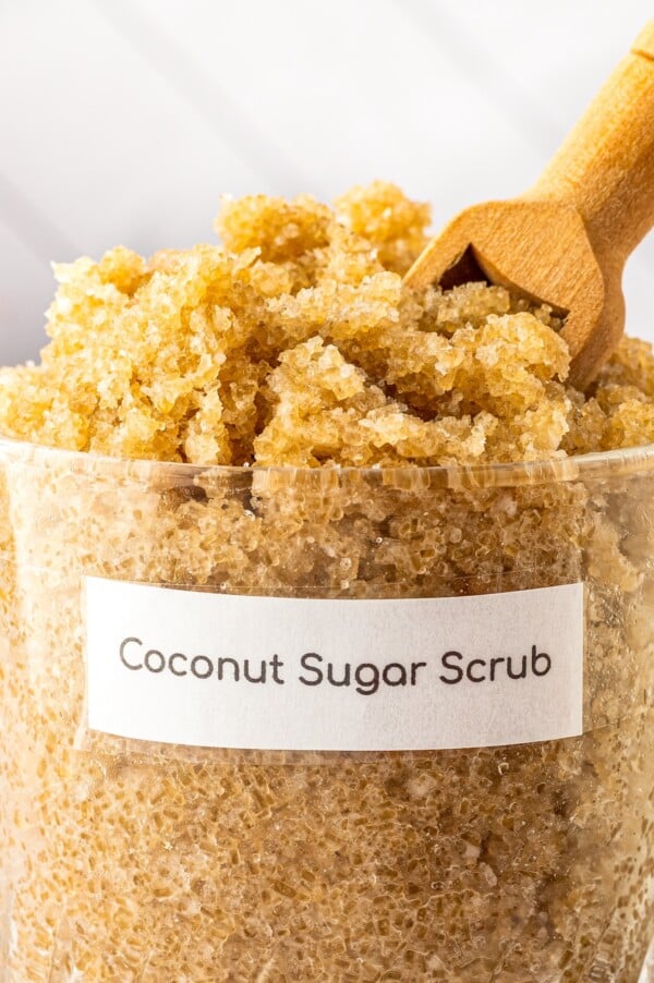 A glass jar filled with diy coconut scrub made with coconut oil and vitamin e oil with a scoop stuck in the top ready to scoop up the sugar body scrub.
