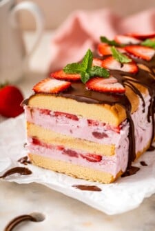 A layered strawberry ice cream cake topped with sliced strawberries, chocolate drizzle, and a mint sprig, set against a blurred background.