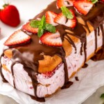 A no-bake strawberry ice cream cake topped with chocolate drizzle, fresh strawberry slices, and mint leaves, with a bowl of whole strawberries and a pink cloth in the background.
