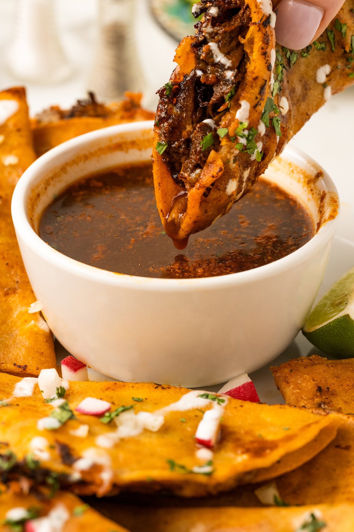 Tacos de birria on a platter and one Birria Tacos being dunked in a bowl of warm birria consommé.