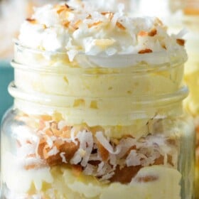 A mason jar with toasted coconut pudding with layers of vanilla wafers, shredded toasted coconut and creamy coconut pudding with whipped cream and more toasted coconut on top.