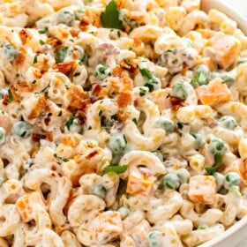 Creamy bacon ranch pasta salad with cheddar cheese and peas in a large white serving bowl with crispy bacon sprinkled on top.