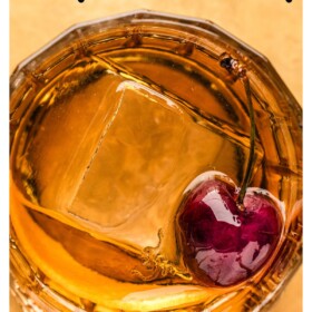 Old fashioned cocktail served in a short glass with a large ice cube ball and a cherry.