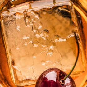 Old fashioned cocktail in a short glass with a large ice cube and a cherry.