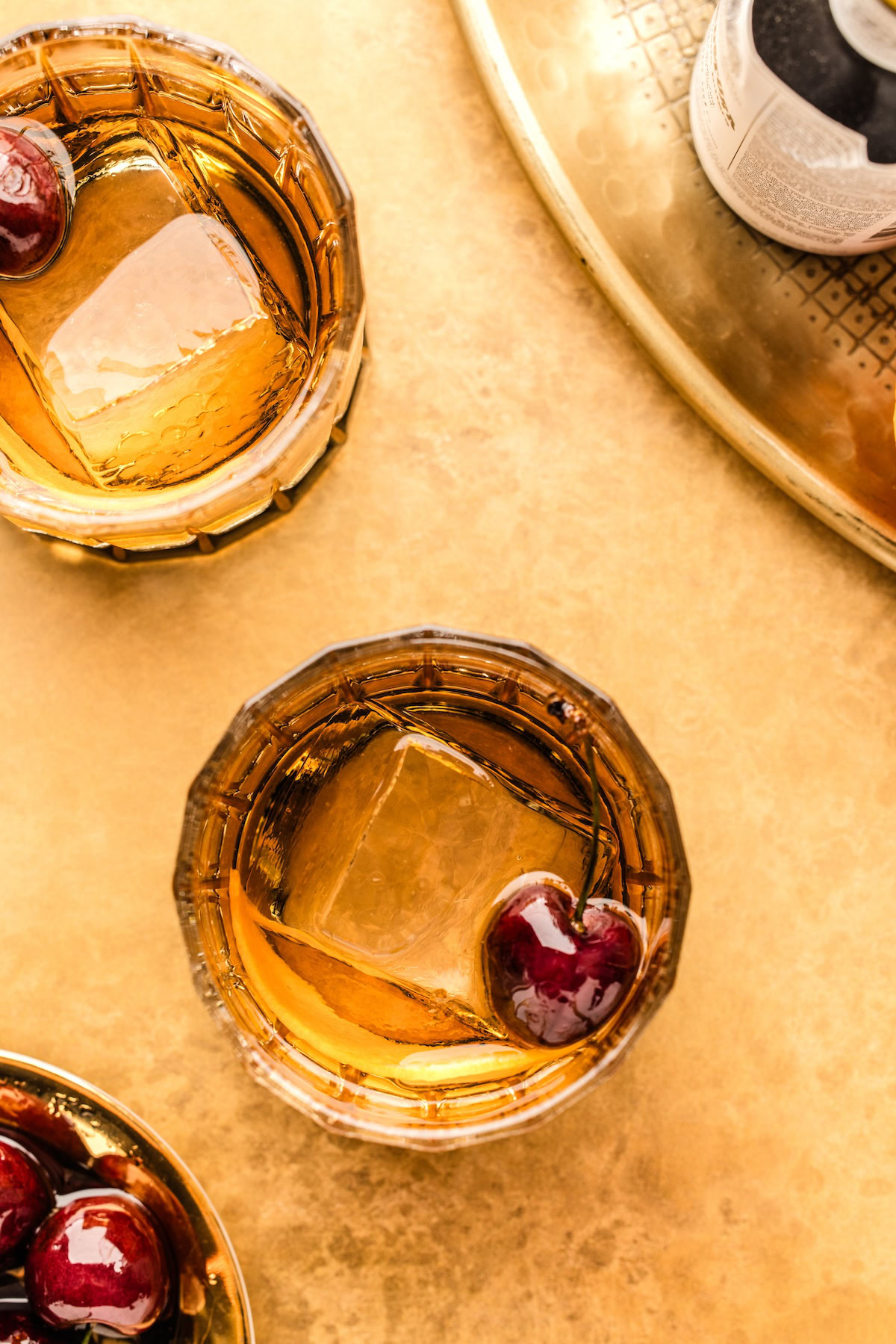 Two old fashioned cocktails garnished with orange peel and cherries next to a bowl of fresh cherries.