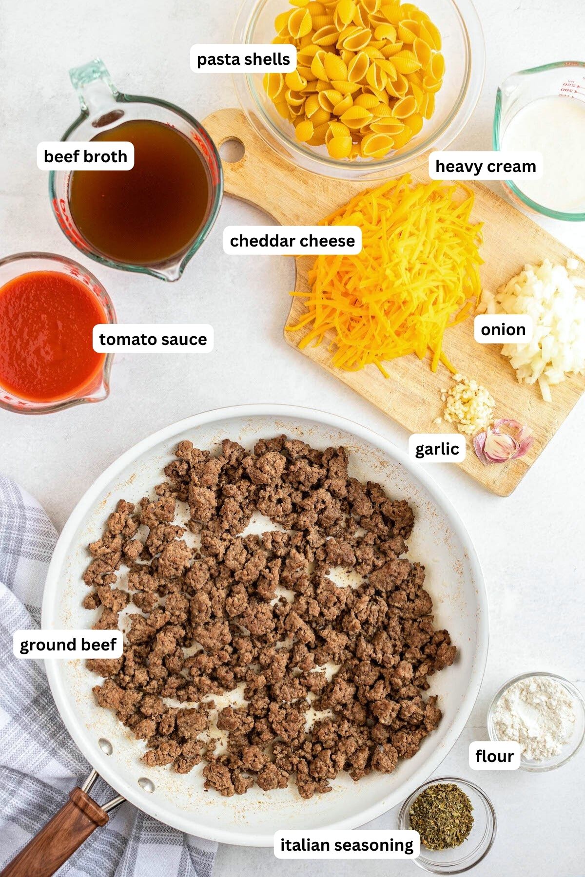 Ingredients arranged in bowls and a skillet for homemade hamburger helper recipe. From top to bottom: pasta shells, beef broth, heavy cream, cheddar cheese, tomato broth, onion, garlic, ground beef, flour and Italian seasoning.