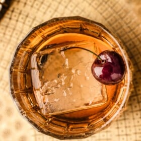 Old fashioned cocktail served in a short glass with a large cube ice cube, orange peel and cherry for garnish.