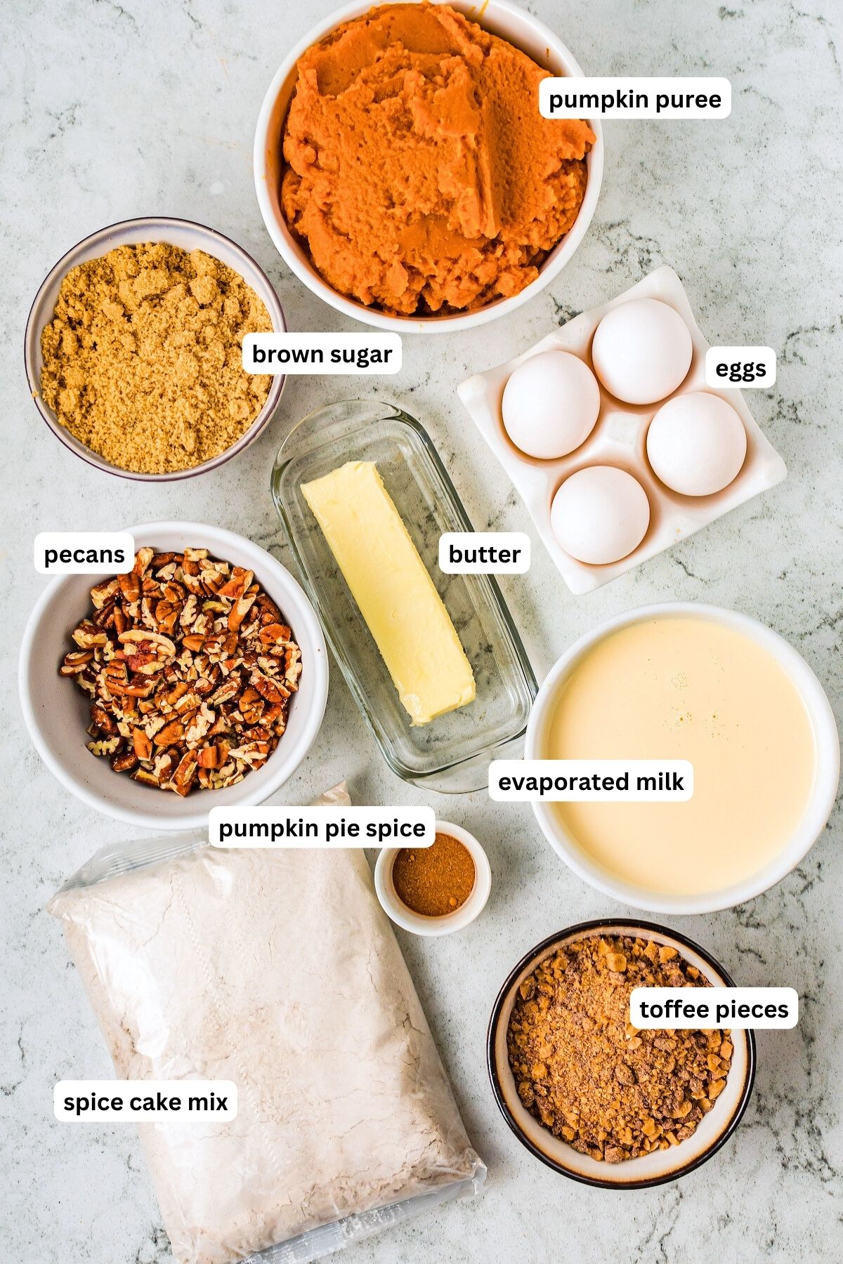 Ingredients in bowls for pumpkin dump cake recipe. From top to bottom: pumpkin puree, brown sugar, eggs, butter, pecans, pumpkin pie spice, evaporated milk, spice cake mix and toffee pieces.