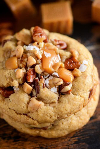 Three salted caramel crunch cookies with pecans and toffee bits stacked on top of each other on a wood cutting board.