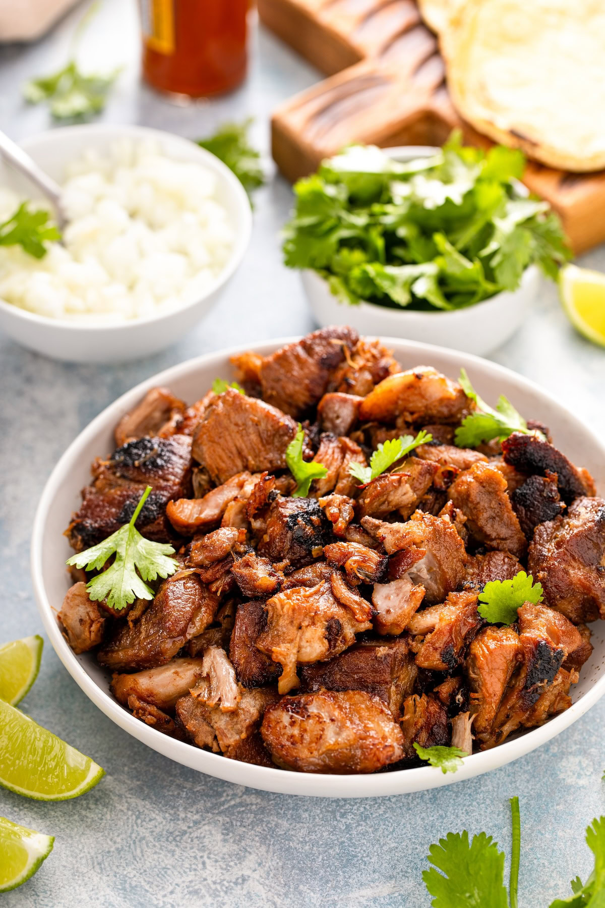 Tender, juicy and crispy pork carnitas in a serving bowl with all the taco fixings behind them in the background.