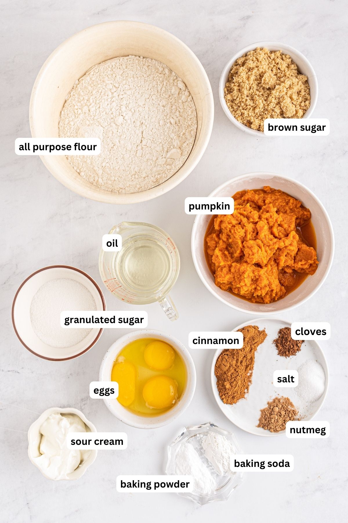 Ingredients arranged in bowls for pumpkin muffin recipe. From top to bottom: All-purpose flour, brown sugar, white sugar, pumpkin, eggs and an extra egg yolk, spices and salt, sour cream, baking powder, baking soda.