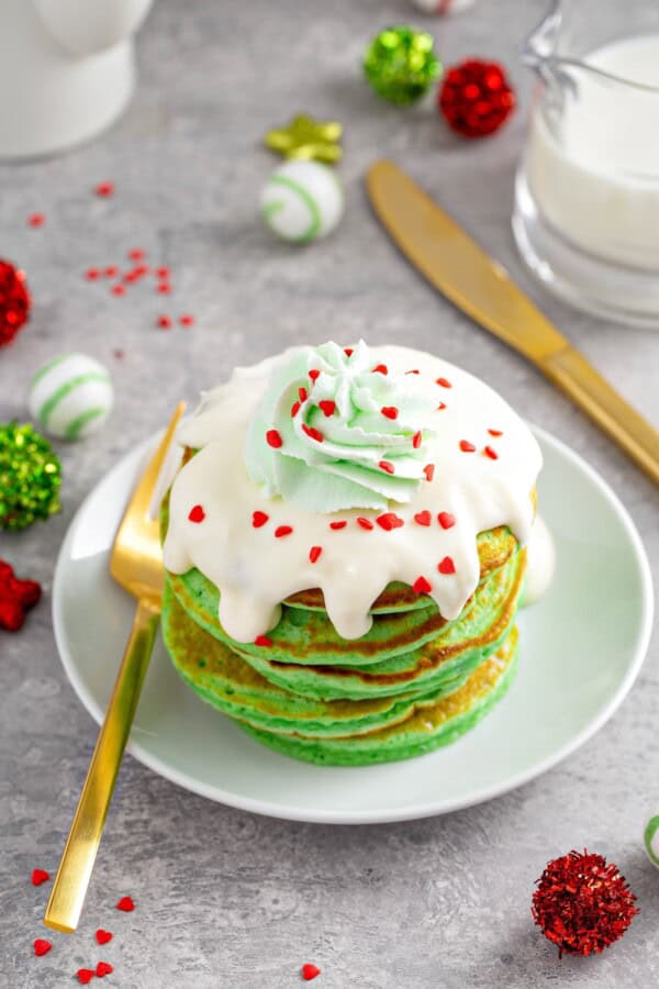 Fluffy green grinch pancakes on a plate with cream cheese frosting, green whipped cream and red heart sprinkles on top.