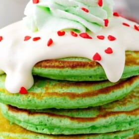 A stack of green Christmas grinch pancakes topped with cream cheese icing, green whipped cream and red heart sprinkles.