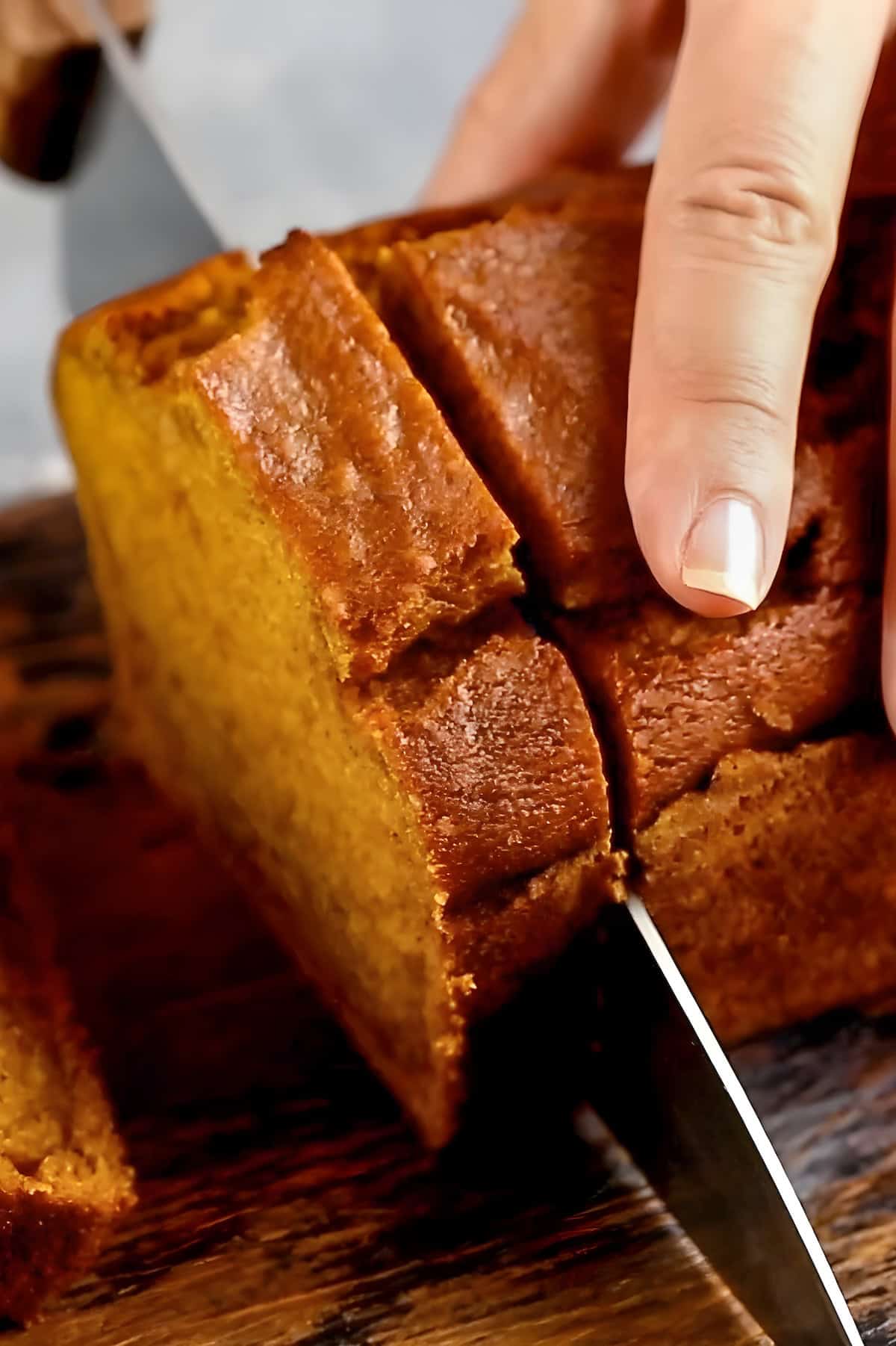 A loaf of moist pumpkin bread is being sliced into pieces with a large knife on a wood cutting board.