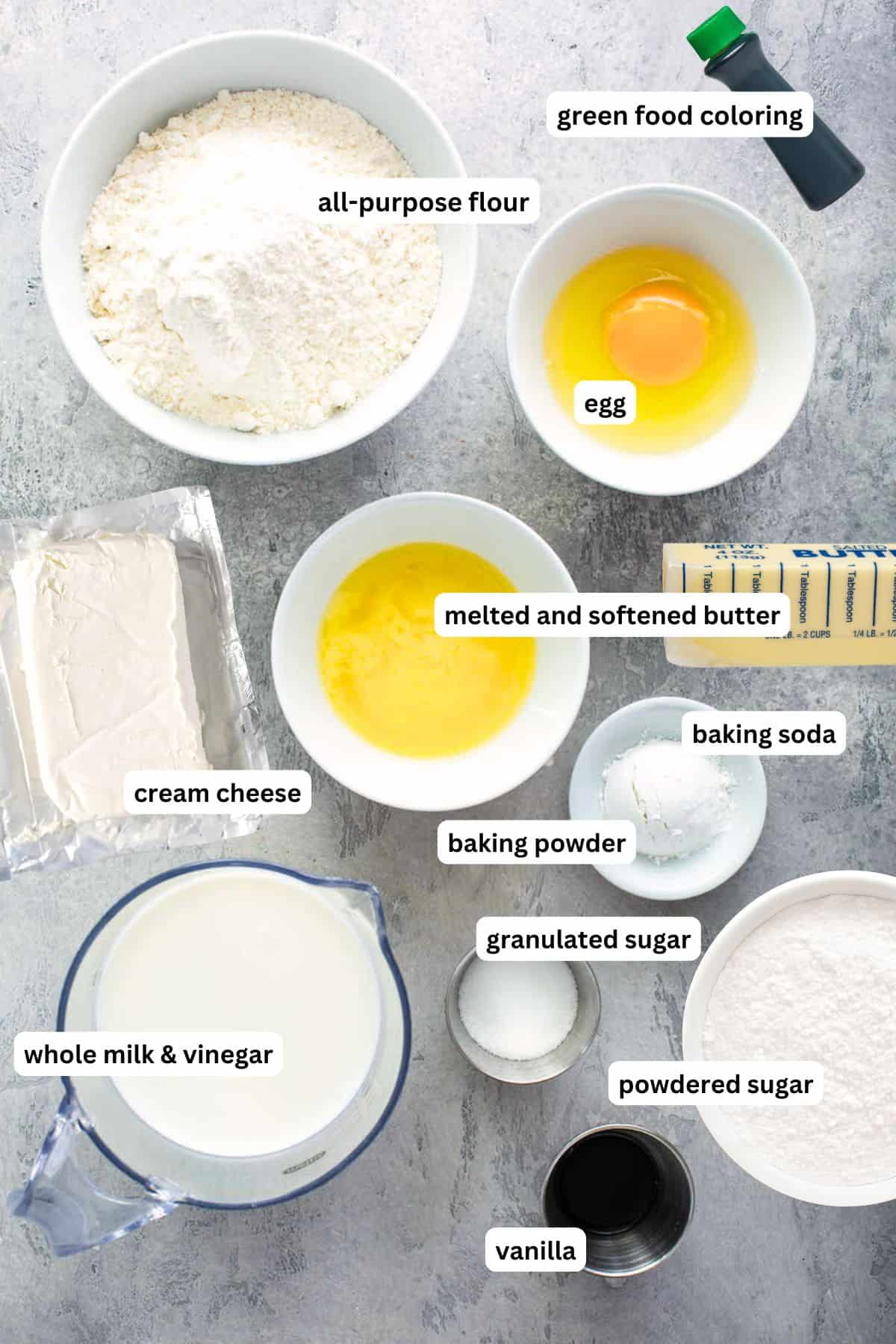 Ingredients for Christmas Grinch pancake recipe arranged in bowls. From top to bottom: green food coloring, all purpose flour, egg, butter, cream cheese, baking soda, baking powder, granulated sugar, whole milk, vinegar, powdered sugar and vanilla.