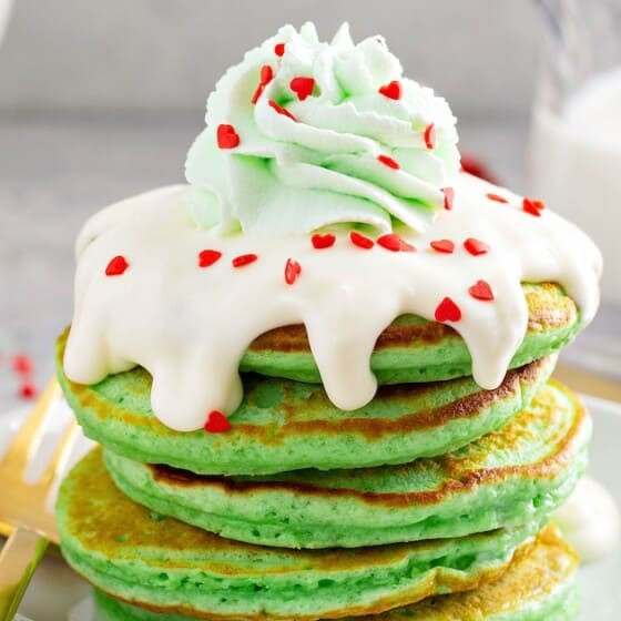 A stick of green Christmas pancakes with cream cheese icing, green whipped cream and red heart sprinkles on top.