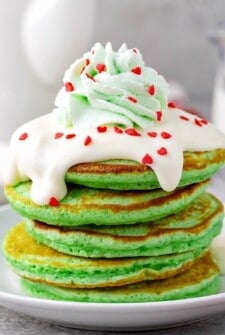 Fluffy stack of green Grinch Christmas pancakes on a plate topped with creamy cream cheese frosting, green tinted whipped cream and red heart sprinkles.