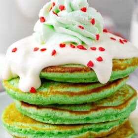 Fluffy stack of green Grinch Christmas pancakes on a plate topped with creamy cream cheese frosting, green tinted whipped cream and red heart sprinkles.