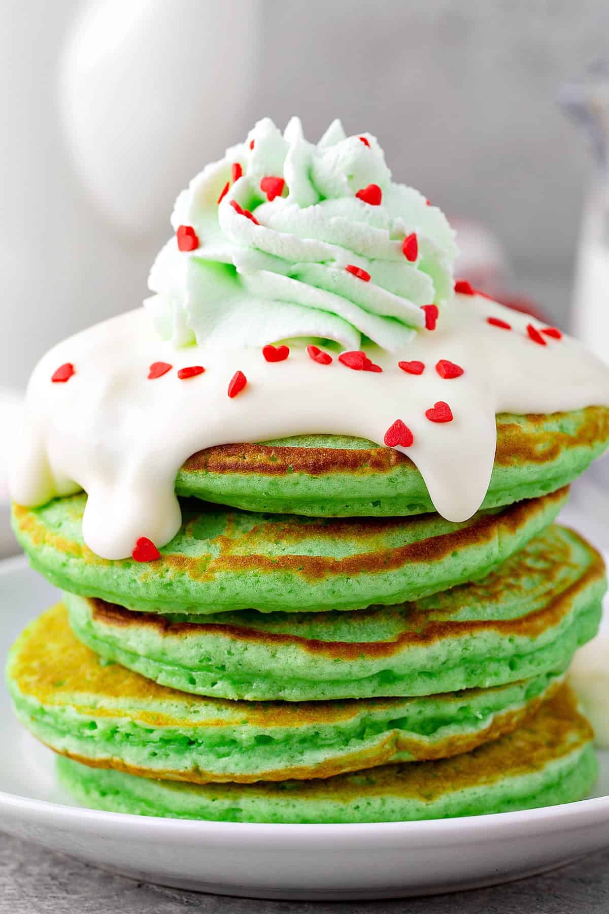 Fluffy stack of green Grinch Christmas pancakes on a plate topped with creamy cream cheese frosting, green tinted whipped cream and red heart sprinkles.