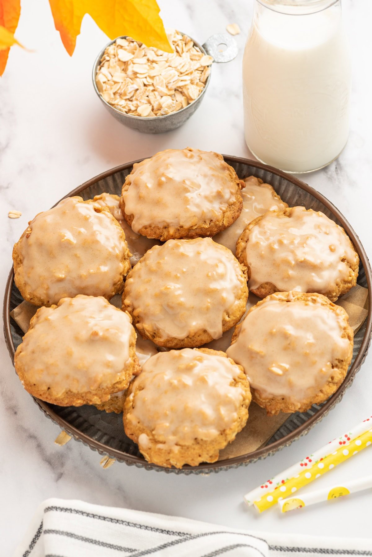 A batch of iced pumpkin oatmeal cookies are presented in a shallow bowl next to a glass of milk and a small cup of dry oats.