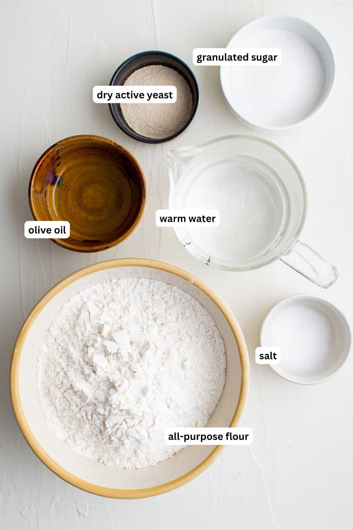 Ingredients for homemade pita bread recipe arranged in bowls. From top to bottom: granulated sugar, dry active yeast, olive oil, warm water, salt and all purpose flour.