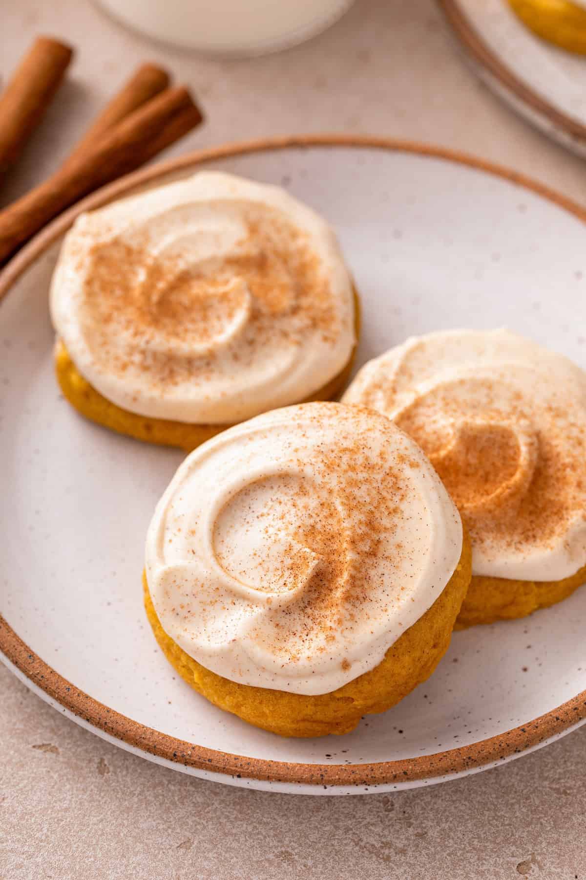 Soft pumpkin cookies with cream cheese icing on a plate next to cinnamon sticks.