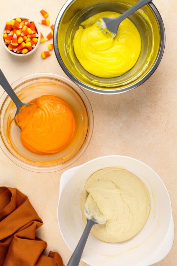 Cake batter is dyed yellow, orange, and white in three different bowls.