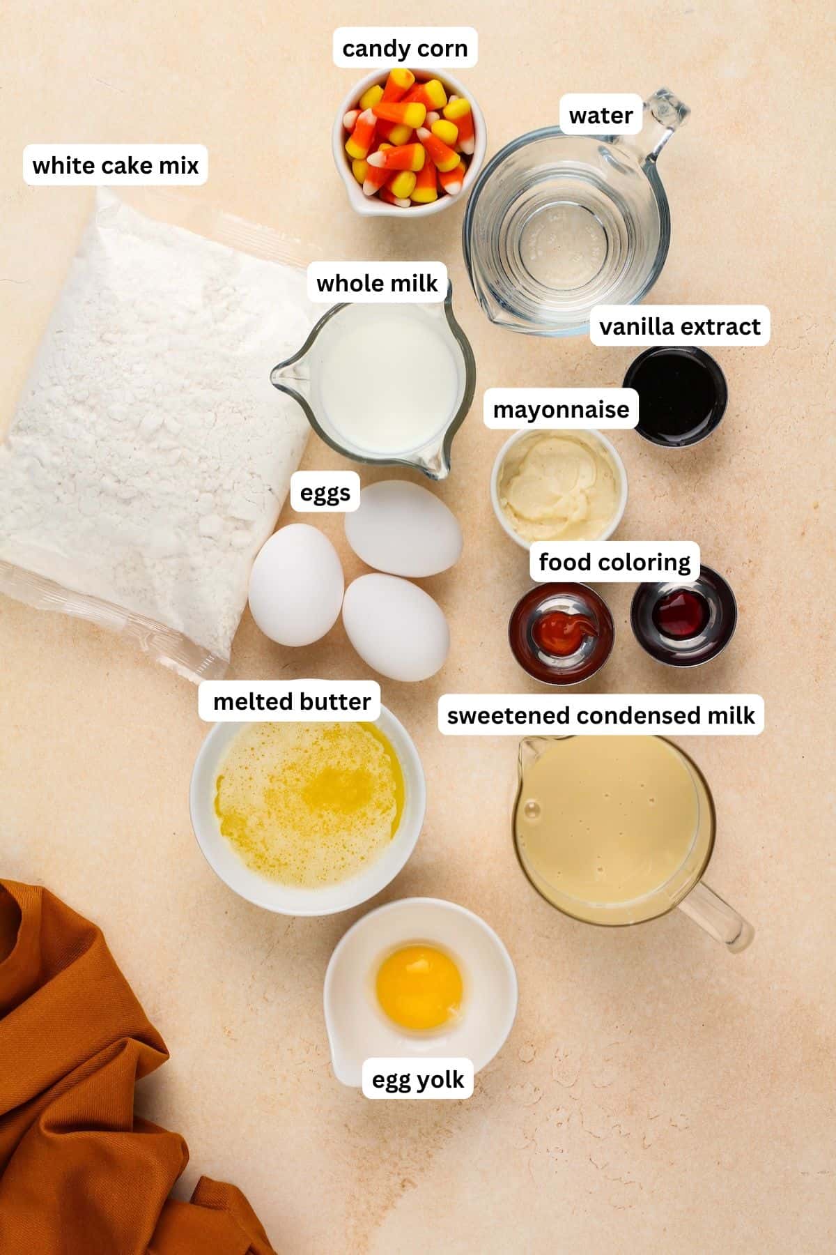 Ingredients for candy corn cake recipe in order from top to bottom: white cake mix, candy corn, water, whole milk, mayonnaise, vanilla extract, eggs, food coloring, melted butter, sweetened condensed milk, egg yolk.