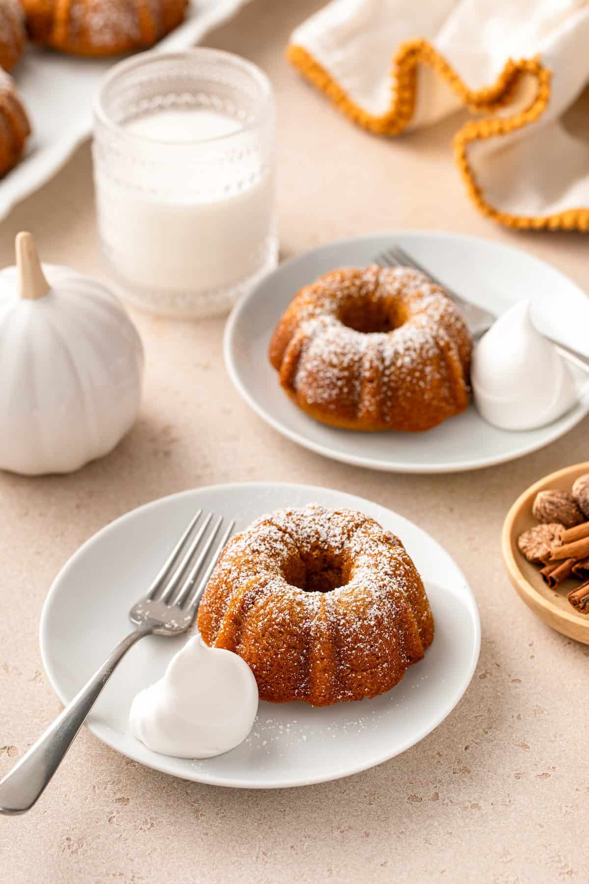 A few white plates are topped with single mini pumpkin bundt cakes with powdered sugar dusted on top and a dollop of whipped cream on the side.